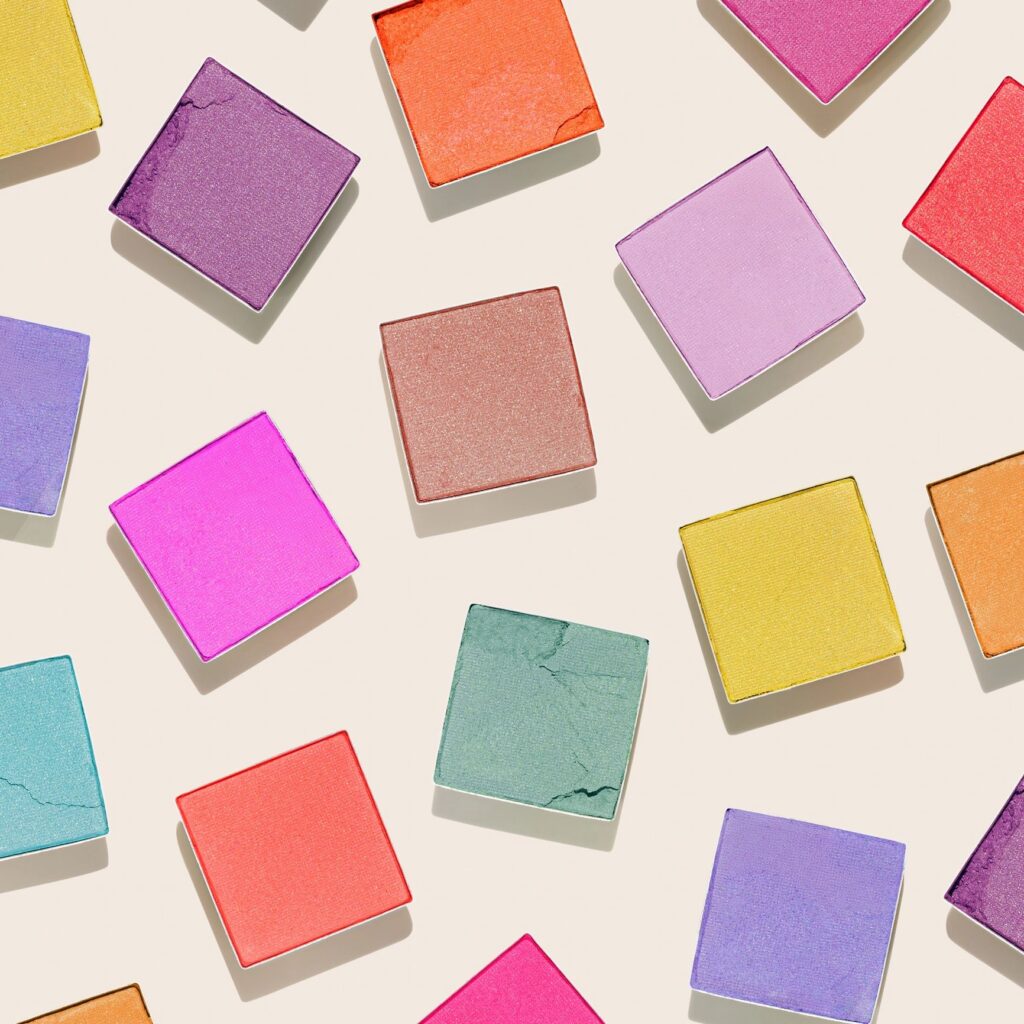 Vivid colored eye shadow makeup palette purple, red, blue, yellow swatches on light beige, flat lay pattern. Woman cosmetics and beauty product concept, collection modern shimmering powder eyeshadows